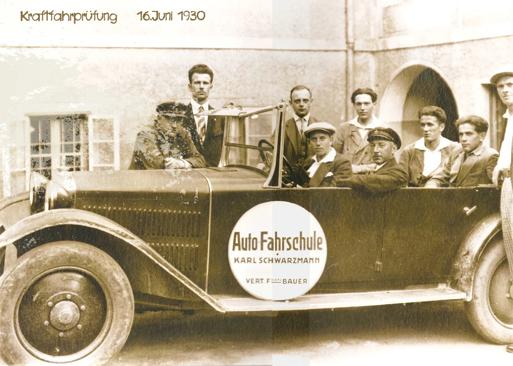 Historisches Foto vom 16. Juni 1930: ein offenes Fahrschulauto mit der Aufschrift „Auto-Fahrschule Karl Schwarzmann“, darin und dahinter Fahrschüler und Prüfer