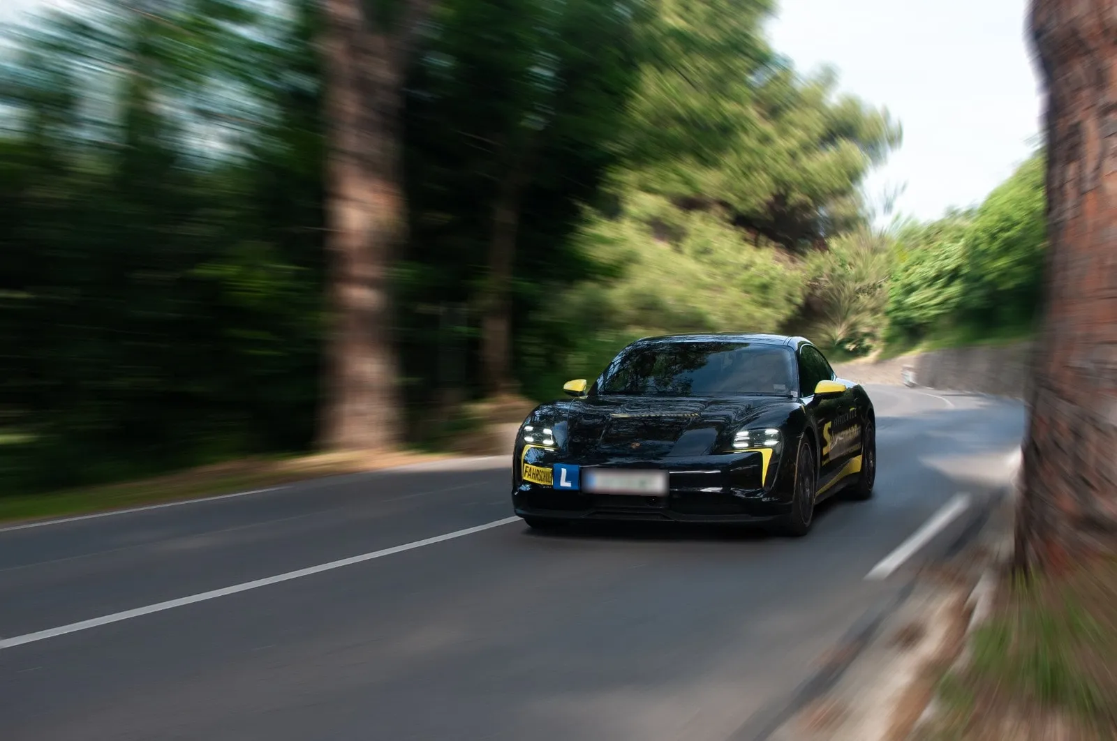 Ein schwarzer Porsche Taycan mit der gelben Beschriftung der Fahrschule Schwarzmann fährt durch eine Waldkurve